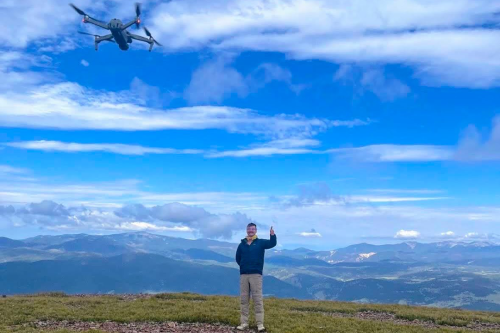 A person waving up at a drone in the sky, against a backdrop of blue sky, clouds and grass