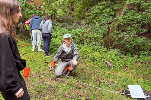 A geography student kneeling in a grassy wood taking a measurement 